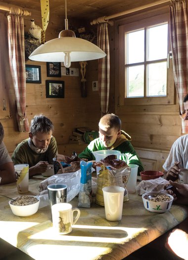 Familie und Kinder sitzen beim Frühstück an einem Holztisch in einer gemütlichen, holzverkleideten Küche, umgeben von Sonnenlicht. | © Kleinwalsertal Tourismus eGen • Fotograf: Andre Tappe