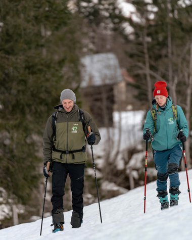 Two people hiking in winter nature through the snow, both with backpacks and trekking poles. | © Kleinwalsertal Tourismus eGen • Photographer: Basti Heckl