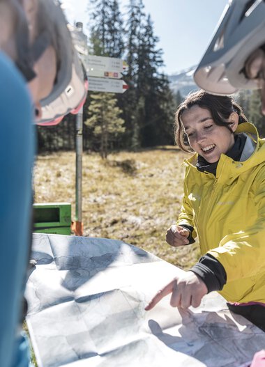 Eine Gruppe von Menschen mit Helmen, die sich um eine Karte beim E-Bike-Trip im Bärguntal versammeln, im Hintergrund sind Bäume und Berge sichtbar. | © Kleinwalsertal Tourismus eGen • Fotograf: Bastian Morell