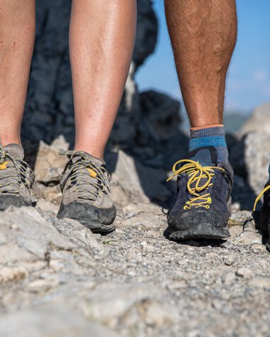 Close-up of three hiking feet on a rocky trail, wearing different colored hiking shoes. | © Kleinwalsertal Tourismus eGen • Photographer: Bastian Morell