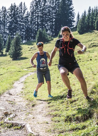 Two trail runners on a forest trail in the mountains on a sunny day, one jumping over an obstacle. | ©   Kleinwalsertal Tourismus eGen • Photographer: Dominik Berchtold