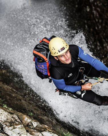 Adventurer rappelling down a waterfall, wearing a helmet and full gear, holding onto a rope in a rugged outdoor setting. | © Kleinwalsertal Tourismus eGen • Photographer: Dominik Berchtold