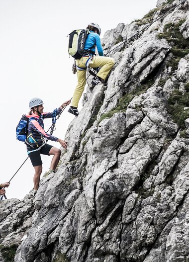 People climbing a steep rock face in the mountains, equipped with climbing gear and helmets. | © Kleinwalsertal Tourismus eGen • Photographer: Dominik Berchtold