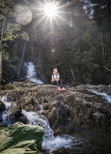 Person sitting on rocks in the Schwarzwasserbach stream in a forest, sunlight shining through trees, water flowing around them. | © Kleinwalsertal Tourismus eGen • Photographer: Dominik Berchtold