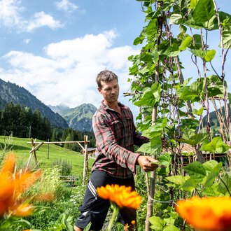 Man in a permaculture garden, surrounded by blooming plants and green mountains in the background, on a sunny day. | © Kleinwalsertal Tourismus eGen • Photographer: Dominik Berchtold