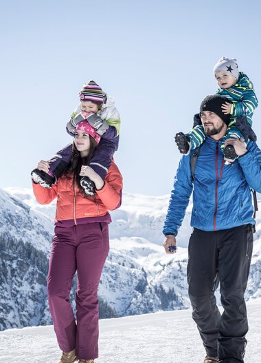 Family winter hiking in the mountains, two children on parents' shoulders, snowy peaks in the background. | © Kleinwalsertal Tourismus eGen • Photographer: Dominik Berchtold