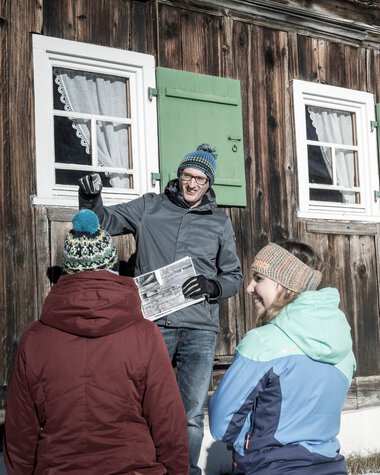 Five people in winter clothing standing in front of an old wooden house, one person holding a newspaper, others listening. | © Kleinwalsertal Tourismus eGen • Photographer: Dominik Berchtold