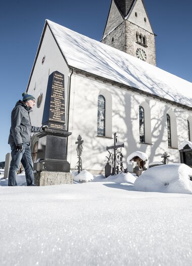 Three people standing in front of an old church in snowy winter with a clear blue sky. | © Kleinwalsertal Tourismus eGen • Photographer: Dominik Berchtold