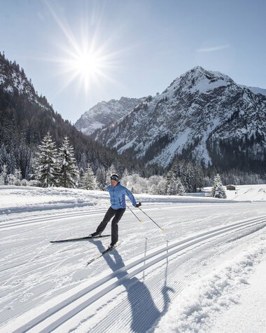 Cross-country skier on a snowy trail in a winter landscape with mountains and the sun in the background. | © Kleinwalsertal Tourismus eGen • Photographer: Dominik Berchtold