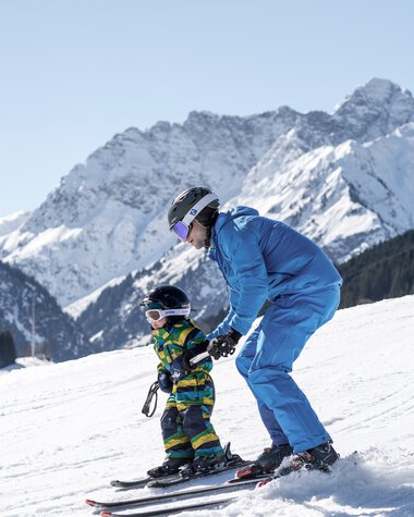 An adult helping a child ski on a snowy slope with mountain peaks in the background. | © Kleinwalsertal Tourismus eGen • Photographer: Dominik Berchtold