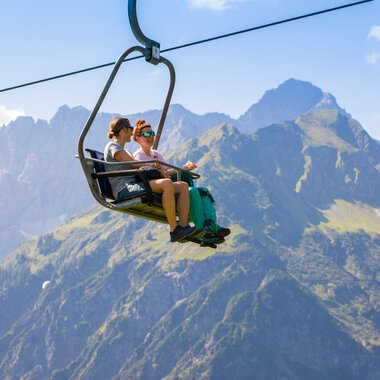 Zwei Personen sitzen in einer Seilbahn über den Bergen des Kleinwalsertals, mit hohen Gipfeln im Hintergrund. | © Kleinwalsertal Tourismus eGen • Fotograf: Frank Drechsel