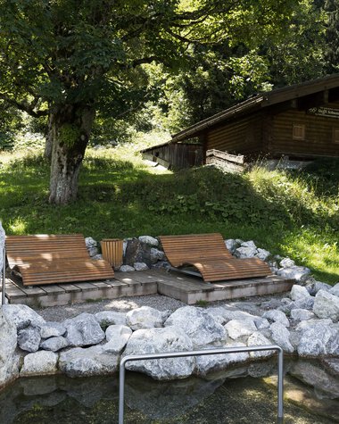 Naturerlebnisplatz mit Wassertretbecken und Steinen, umgeben von Bäumen und einem kleinen Holzhaus im Hintergrund. | © Kleinwalsertal Tourismus eGen • Fotograf: Oliver Farys