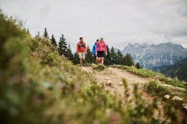 Drei Wanderer auf einer Bergwanderung im Kleinwalsertal, umgeben von grünen Wiesen und Bergen, bei bedecktem Himmel. | © Kleinwalsertal Tourismus eGen • Fotograf: Oliver Farys