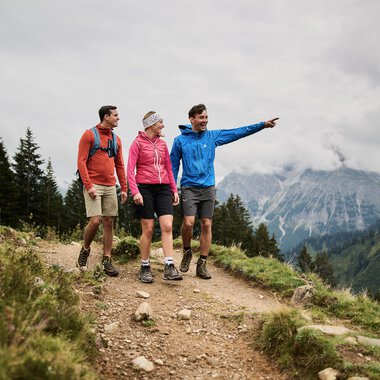 Vier Wanderer, darunter zwei Frauen und zwei Männer, wandern auf einem Bergpfad in den Bergen, einer der Männer zeigt nach vorne. | © Kleinwalsertal Tourismus eGen • Fotograf: Oliver Farys