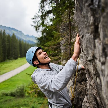 Junge Frau beim Klettern im Klettergarten, trägt Helm und Sicherheitsausrüstung, umgeben von Bergen und Wäldern. | © Kleinwalsertal Tourismus eGen • Fotograf: Oliver Farys