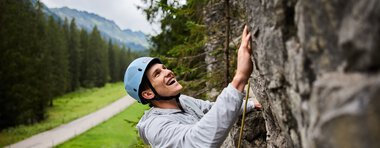 Young woman climbing in a rock climbing area, wearing a helmet and safety gear, with mountains and forests in the background. | © Kleinwalsertal Tourismus eGen • Photographer: Oliver Farys