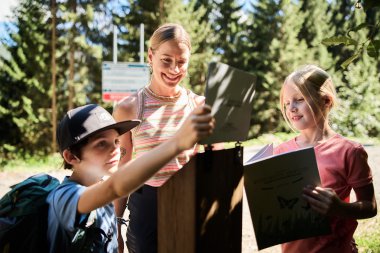 Three children and a young woman engaging in a nature explorer adventure in the mountains, all smiling and looking at a tablet and a brochure. | © Kleinwalsertal Tourismus eGen • Photographer: Oliver Farys