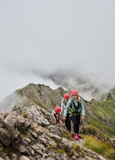 People on a via ferrata in the mountains surrounded by clouds and rocks, with Walser Mountain School logo visible. | © Kleinwalsertal Tourismus eGen • Photographer: Oliver Farys