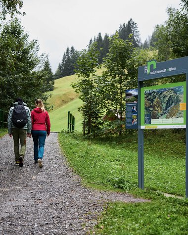 Two people hiking on a forest trail in Kleinwalsertal, passing informational signs and surrounded by trees. | © Kleinwalsertal Tourismus eGen • Photographer: Oliver Farys