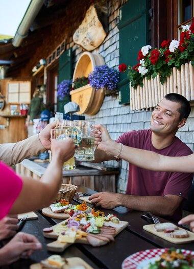 People enjoying summer outdoor dining on a terrasse with wooden houses and colorful flowers, sharing toasts and snacks. | © Kleinwalsertal Tourismus eGen • Photographer: Oliver Farys