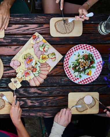 Top-down view of a table with cheese, meat, bread, and salad plates, surrounded by four people enjoying a meal. | © Kleinwalsertal Tourismus eGen • Photographer: Oliver Farys