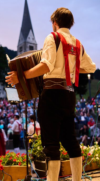 Musician on stage at the Alpenzauber Festival in Kleinwalsertal, playing guitar while the crowd watches, with mountains and a church in the background. | © Kleinwalsertal Tourismus eGen • Photographer: Oliver Farys