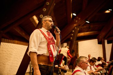 Man in traditional attire singing and performing music at a summer event of the Trachtenkapelle Mittelberg in a wooden hall. | © Kleinwalsertal Tourismus eGen • Photographer: Oliver Farys
