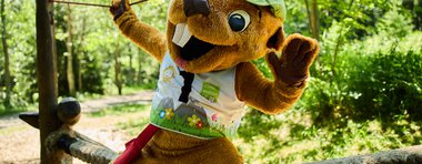 A mascot character in a forest during a summer 2024 event, wearing a green hat and colorful vest, balancing on a rope. | © Kleinwalsertal Tourismus eGen • Photographer: Oliver Farys