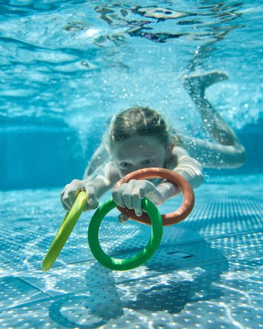 Ein junges Mädchen schwimmt im Freibad, hält sich an bunten Wasserhölzern und schaut nach vorne, während sie unter Wasser ist. | © Kleinwalsertal Tourismus eGen • Fotograf: Oliver Farys