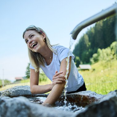 Woman sitting at the Kneipp basin, laughing and enjoying cold water therapy outdoors on a sunny day. | © Kleinwalsertal Tourismus eGen • Photographer: Oliver Farys