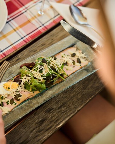 Person schneidet eine kalte Vorspeise mit Lachs, Salat und Samen auf einem Holztablett bei einer kulinarischen Wanderung im Kleinwalsertal. | ©  Kleinwalsertal Tourismus eGen • Fotograf: Oliver Farys