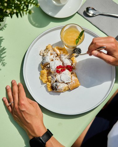 Tisch mit einem Teller mit köstlichem Kaiserschmarrn, garnierte mit Puderzucker und Kirschen, daneben ein Glas frisch gepresster Orangensaft. | © Kleinwalsertal Tourismus eGen • Fotograf: Oliver Farys