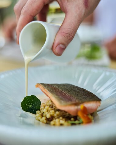 Ein Koch gießt cremige Sauce über ein Gericht mit Fisch, serviert auf einem weißen Teller bei einem kulinarischen Event. | © Kleinwalsertal Tourismus eGen • Fotograf: Oliver Farys