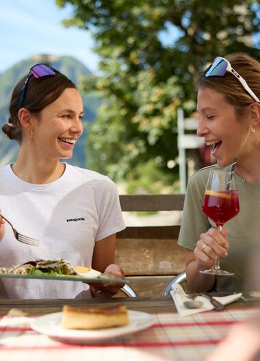 Two smiling women enjoy a mountain picnic outdoors with a scenic mountain view in the background on a summer day. | © Kleinwalsertal Tourismus eGen • Photographer: Oliver Farys