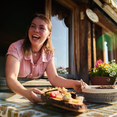 Eine lachende junge Frau steht an einem Fenster der almähnlichen Hütte, hält eine servierte Mahlzeit und genießt die Sommeratmosphäre im Kleinwalsertal. | © Kleinwalsertal Tourismus eGen • Fotograf: Oliver Farys