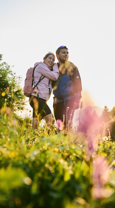 Ein Paar wandert in einem blühenden Feld bei Sonnenaufgang im Kleinwalsertal, umgeben von Natur und Bäumen. | © Kleinwalsertal Tourismus eGen • Fotograf: Oliver Farys