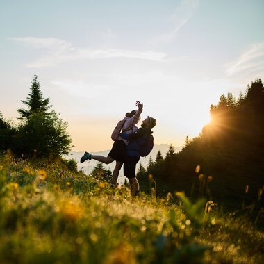 Ein Paar hebt sich bei Sonnenaufgang in einer grünen Bergwiese im Kleinwalsertal hoch, umgeben von Bäumen, mit Sonnenlicht im Hintergrund. | © Kleinwalsertal Tourismus eGen • Fotograf: Oliver Farys