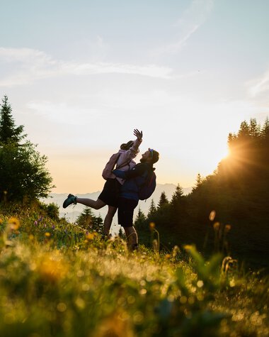A couple lifts each other up on a green mountain meadow at sunrise in Kleinwalsertal, surrounded by trees, with sunlight in the background. | © Kleinwalsertal Tourismus eGen • Photographer: Oliver Farys