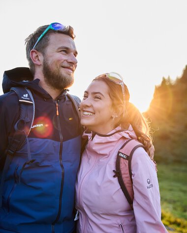 Ein lachendes Paar beim Wandern im Sommer, Sonnenaufgang im Hintergrund in einem Waldgebiet. | © Kleinwalsertal Tourismus eGen • Fotograf: Oliver Farys