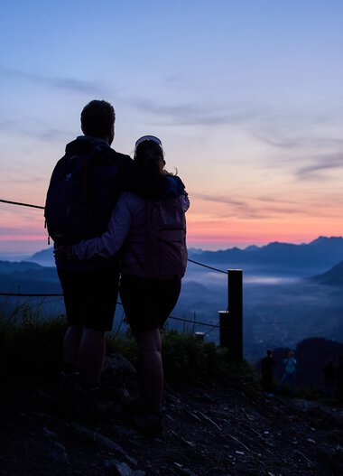 Ein Paar schaut beim Sonnenaufgang vom Aussichtspunkt im Kleinwalsertal auf die Berglandschaft. | © Kleinwalsertal Tourismus eGen • Fotograf: Oliver Farys