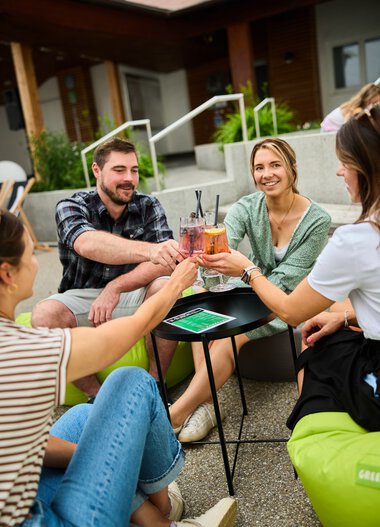 Vier junge Erwachsene sitzen im Sommer vor einem Haus, stoßen mit Getränken an und lachen, während sie eine entspannte Zeit im Freien genießen. | © Kleinwalsertal Tourismus eGen • Fotograf: Oliver Farys