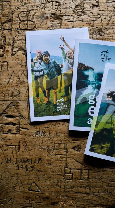 Drei Prospektfächer mit Fotos und Informationen über das Kleinwalsertal, liegen auf einer Holzoberfläche. | © Kleinwalsertal Tourismus eGen • Fotograf: Oliver Farys
