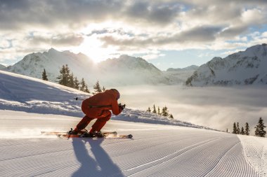 A skier in red gear skiing on a groomed slope in the mountains with sunlight and clouds in the sky. | © Kleinwalsertal Tourismus eGen • Photographer: Oliver Farys