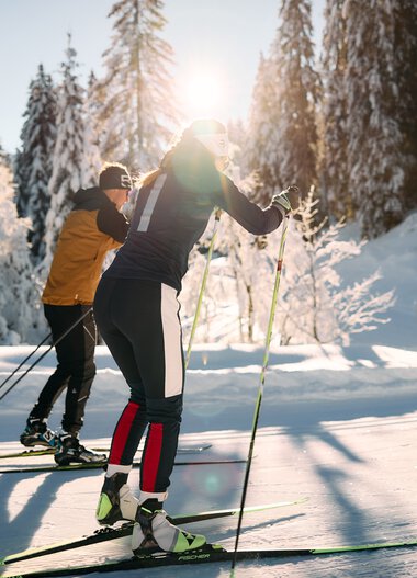 Two cross-country skiers in a snowy winter landscape, sunlight filtering through trees, alpine scenery in the background. | © Kleinwalsertal Tourismus eGen • Photographer: Oliver Farys