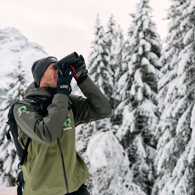 Man in winter clothing with binoculars in snowy forest in the Alps, likely a ranger or nature guide. | © Kleinwalsertal Tourismus eGen • Photographer: Oliver Farys