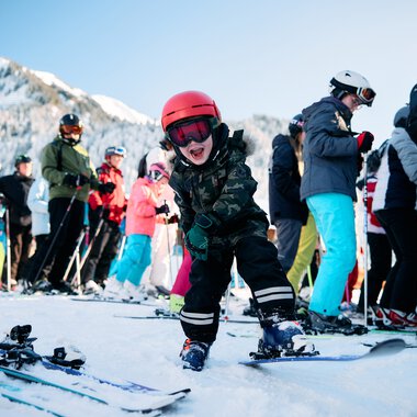 A young boy skiing and smiling, surrounded by other skiers in a snowy mountain landscape. | © Kleinwalsertal Tourismus eGen • Photographer: Oliver Farys