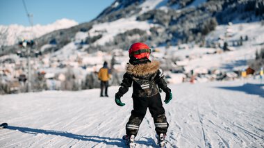 Ein Kind beim Skifahren auf einer Piste im Schnee, umgeben von Bergen und anderen Skifahrern, in winterlicher Landschaft. | © Kleinwalsertal Tourismus eGen • Fotograf: Oliver Farys