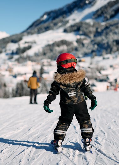 A child skier in winter clothing on a snowy slope, with mountains and other skiers in the background. | © Kleinwalsertal Tourismus eGen • Photographer: Oliver Farys