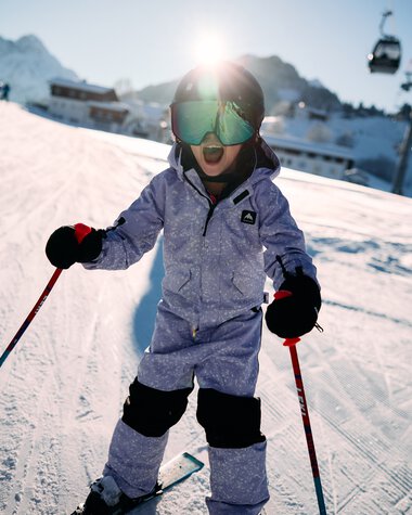 A young child skiing on a slope at a ski resort in Kleinwalsertal, surrounded by mountains, wearing goggles and winter clothing, holding ski poles. | © Kleinwalsertal Tourismus eGen • Photographer: Oliver Farys