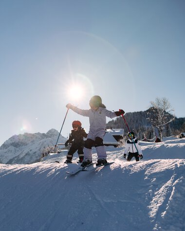 Children skiing on a snow-covered slope in the mountains under sunlight, accompanied by an adult. | © Kleinwalsertal Tourismus eGen • Photographer: Oliver Farys